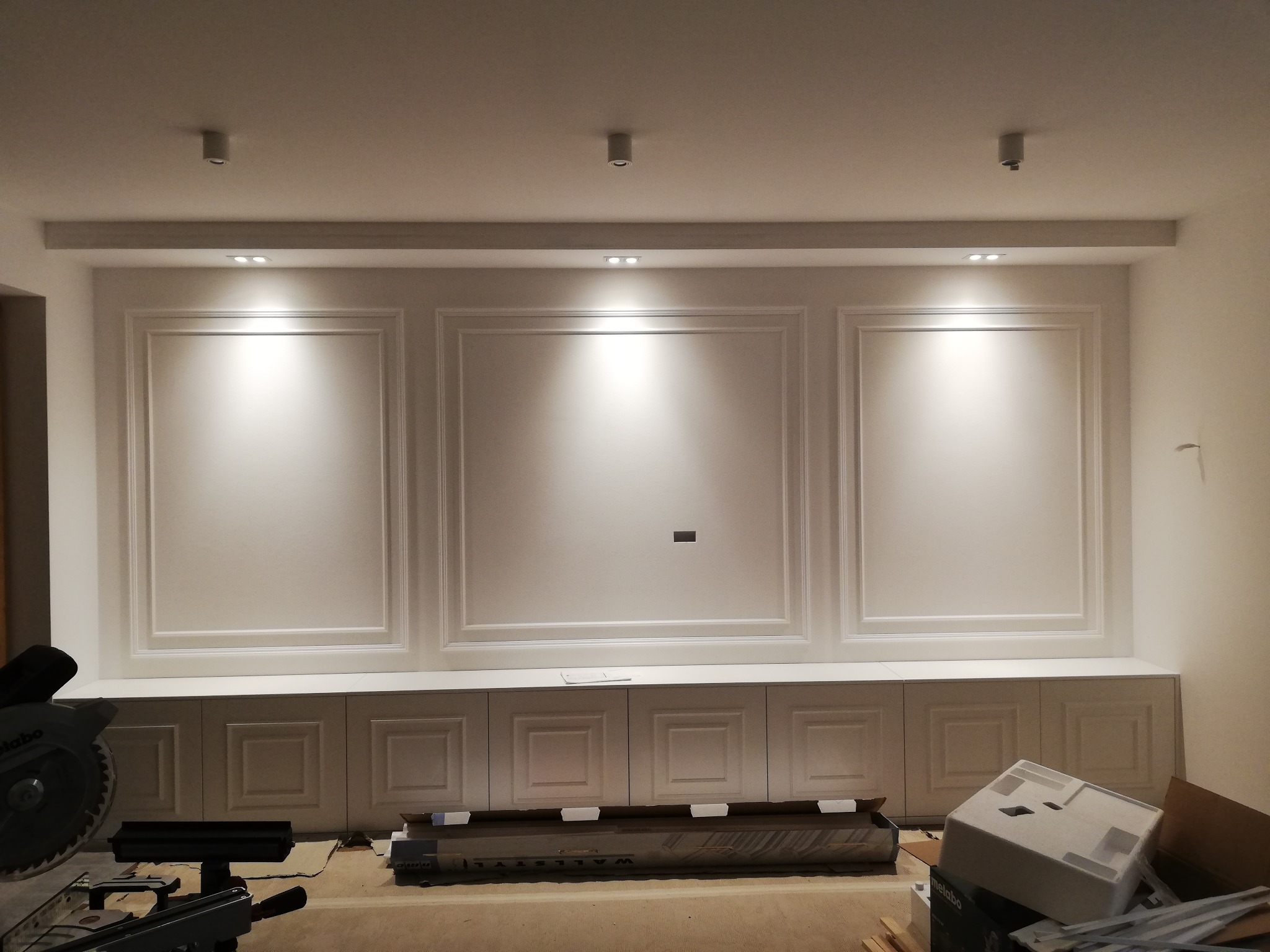 Ściana z dekoracyjnymi panelami w stylu klasycznym i szafkami pod spodem, oświetlona punktowo, z widoczną piłą tarczową i materiałami budowlanymi na podłodze.