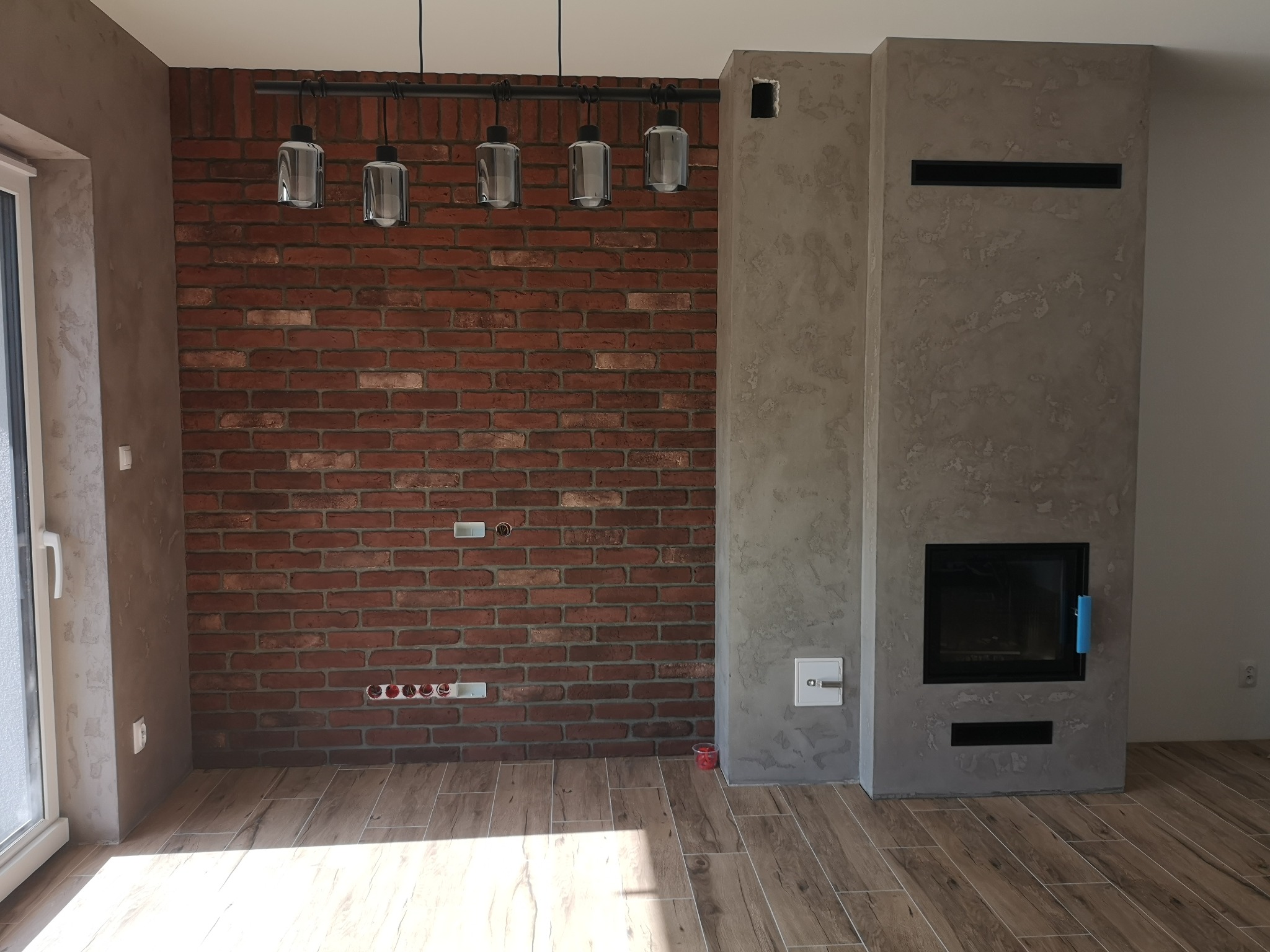 Pokój z ceglaną ścianą, betonowym kominkiem i drewnopodobną podłogą, oświetlony wiszącymi lampami w stylu industrialnym.