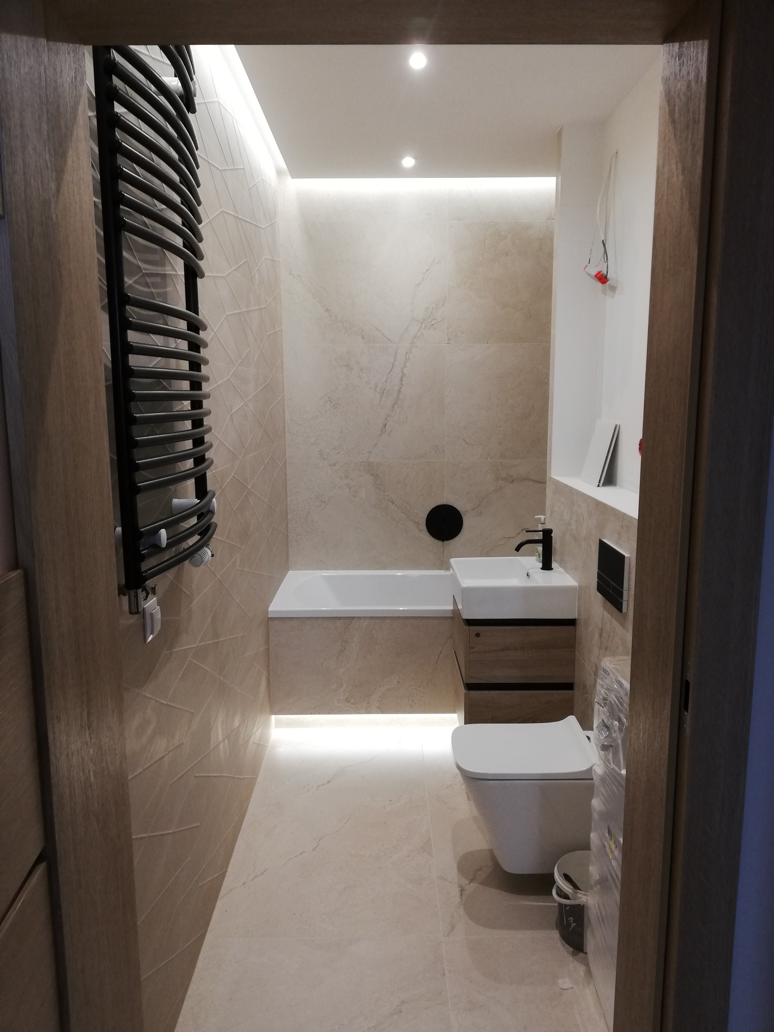 Nowoczesna łazienka z beżowymi płytkami imitującymi marmur, podświetleniem LED pod wanną i nad lustrem, wiszącą toaletą i grzejnikiem drabinkowym w kolorze czarnym.