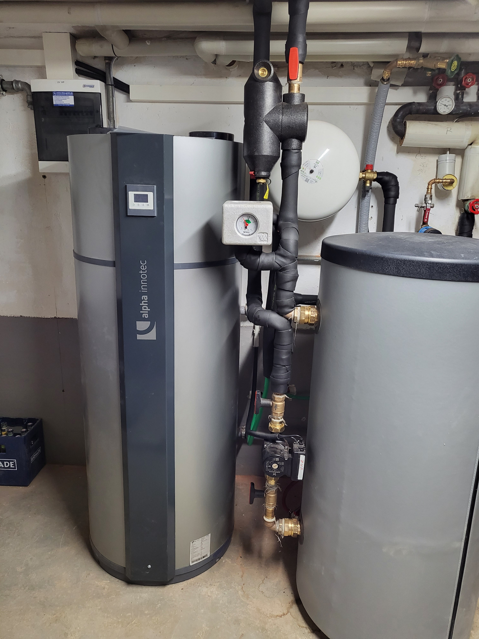 Dwa szare zbiorniki, jeden z logo Alpha Innotec, połączone rurami z izolacją, manometr na rurze, instalacja w piwnicy.