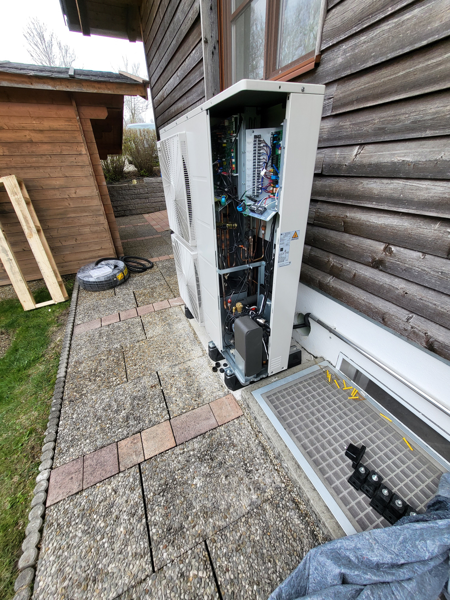 Otwarty panel boczny pompy ciepła, ukazujący wewnętrzne okablowanie i komponenty, zainstalowanej przy drewnianym budynku we Wrocławiu, z widocznymi elementami instalacyjnymi obok.