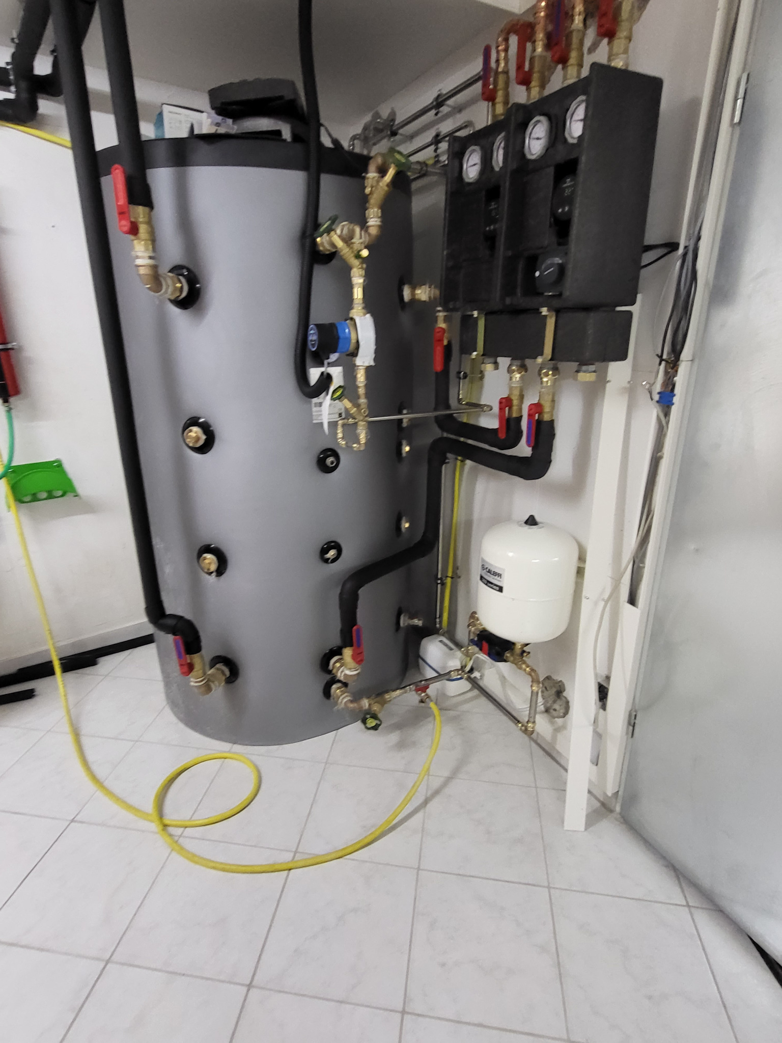 Szary zbiornik buforowy z czarnymi rurami izolowanymi, manometry i biały zbiornik przeponowy Calpeda, instalacja grzewcza w kotłowni na tle białych płytek.