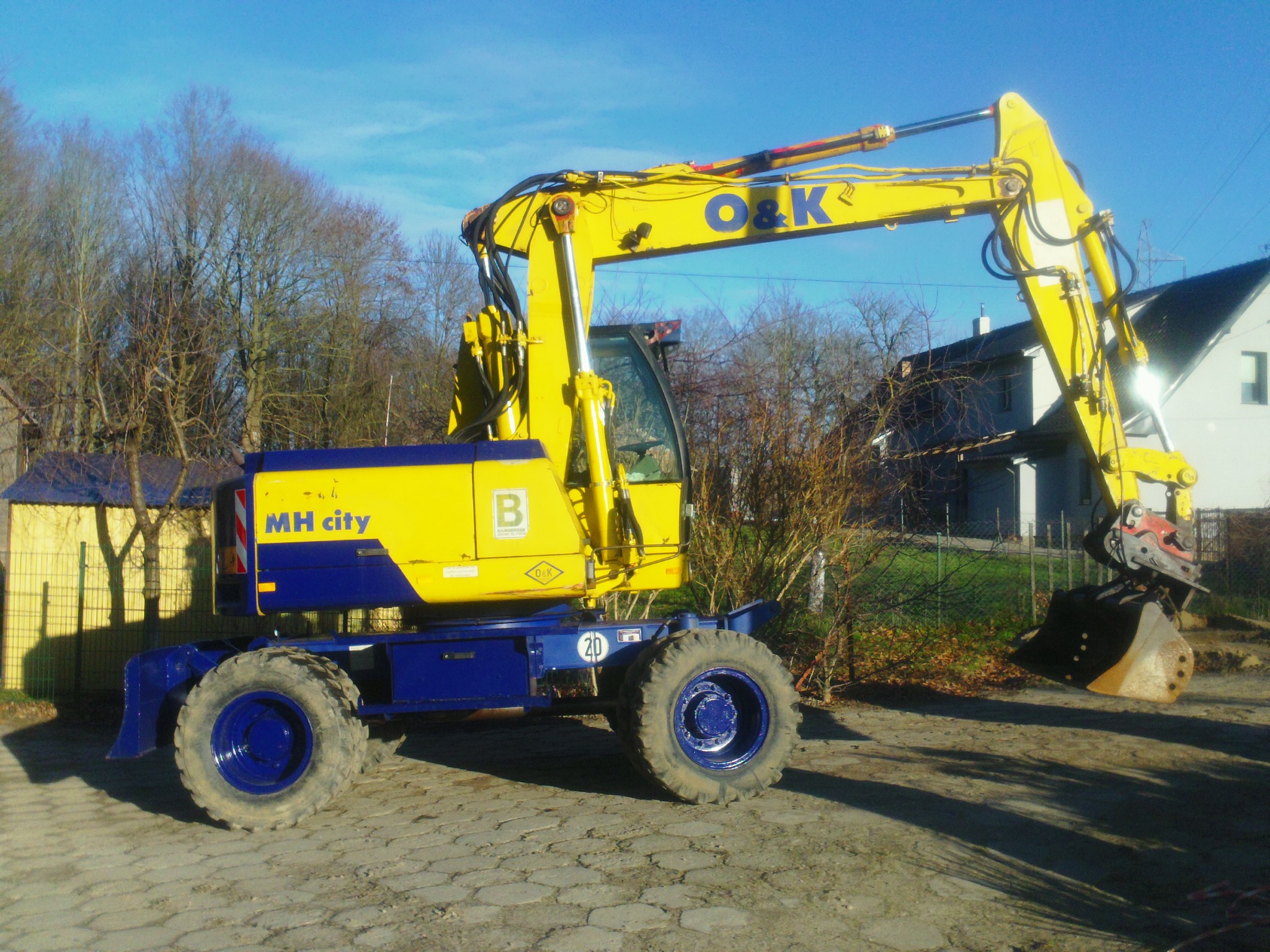 Żółto-niebieska koparka O&K MH City na brukowanej powierzchni, z domem i drzewami w tle, w słoneczny dzień. Widoczny numer 20 na maszynie.