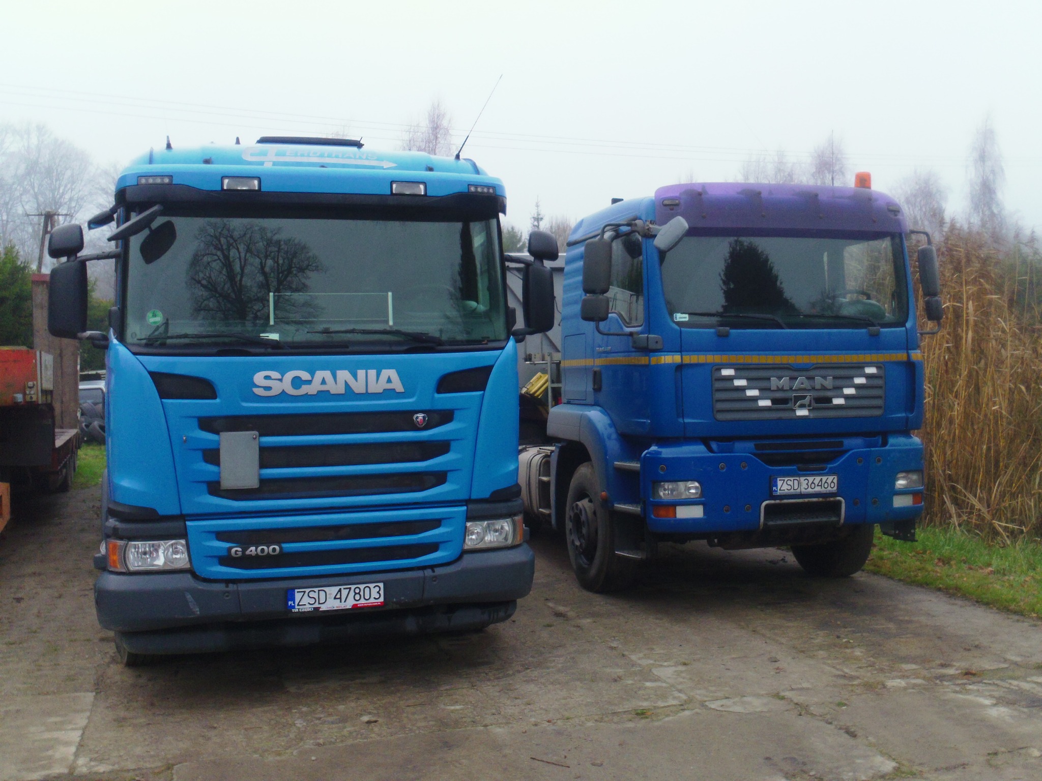 Dwa niebieskie samochody ciężarowe: Scania G400 i MAN, stojące obok siebie na betonowej nawierzchni. Widoczne tablice rejestracyjne i detale pojazdów.