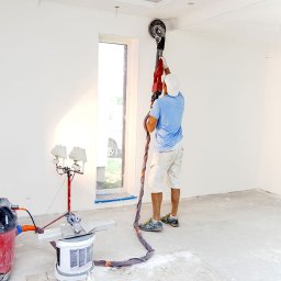 MichtMar - Pracownik w Gościcino szlifuje ścianę szlifierką z odkurzaczem, przygotowując ją do dalszych prac wykończeniowych. Widoczne dodatkowe oświetlenie na statywie.