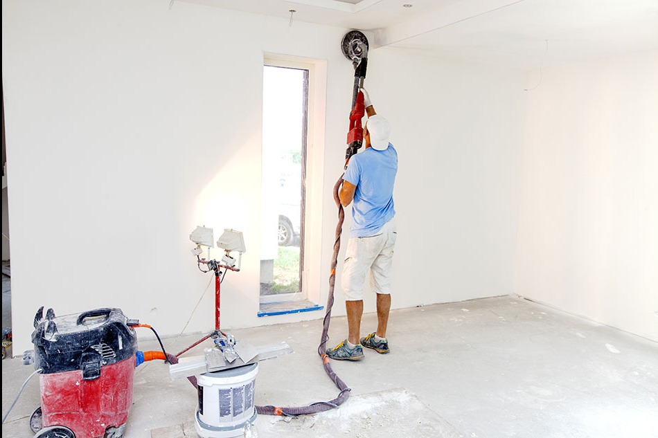 Pracownik w Gościcino szlifuje ścianę szlifierką z odkurzaczem, przygotowując ją do dalszych prac wykończeniowych. Widoczne dodatkowe oświetlenie na statywie.