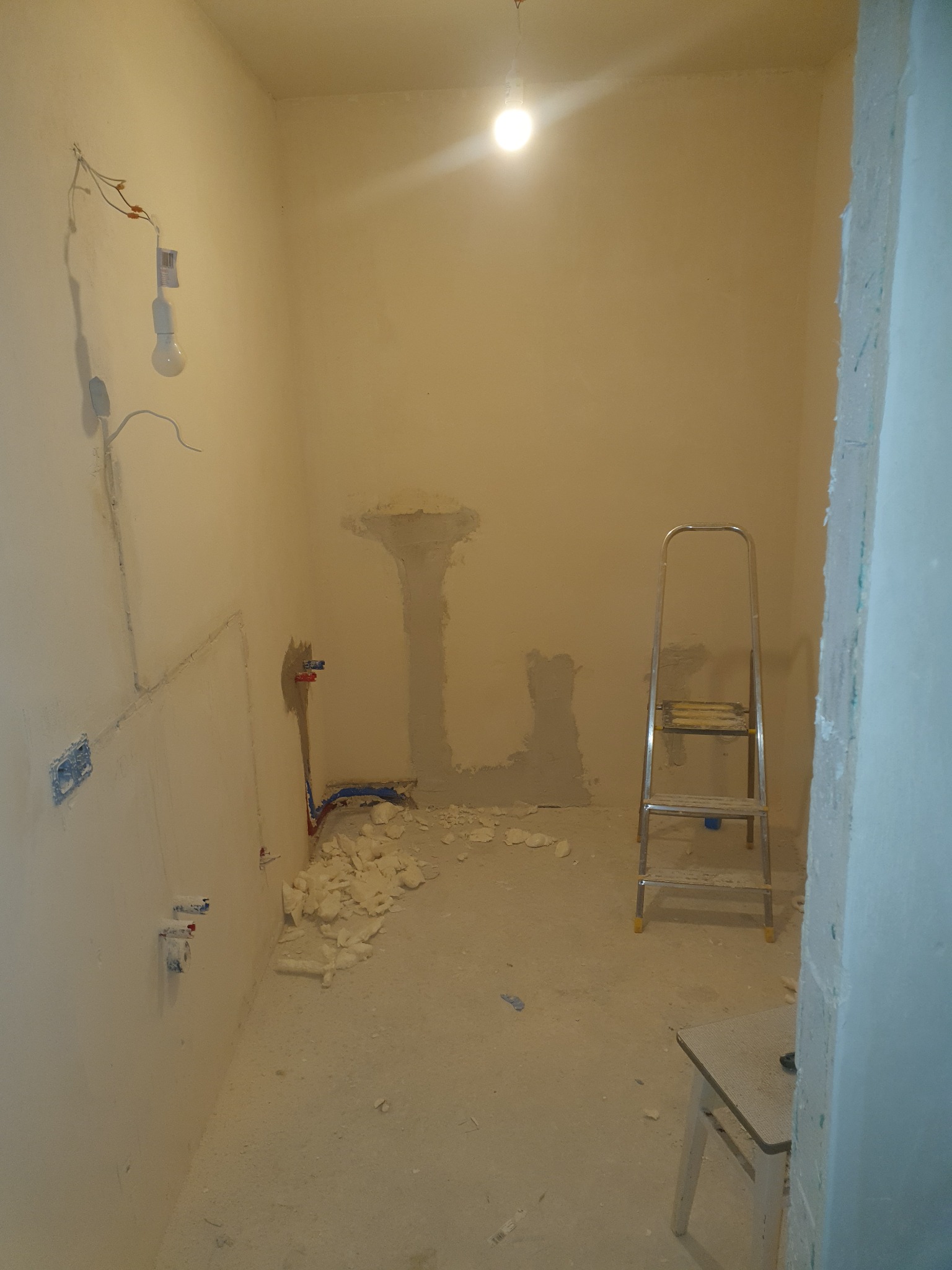 Wnętrze pomieszczenia w trakcie prac przygotowawczych: widoczne przewody elektryczne, rury hydrauliczne, fragmenty gładzi na ścianach, drabina aluminiowa i krzesło.
