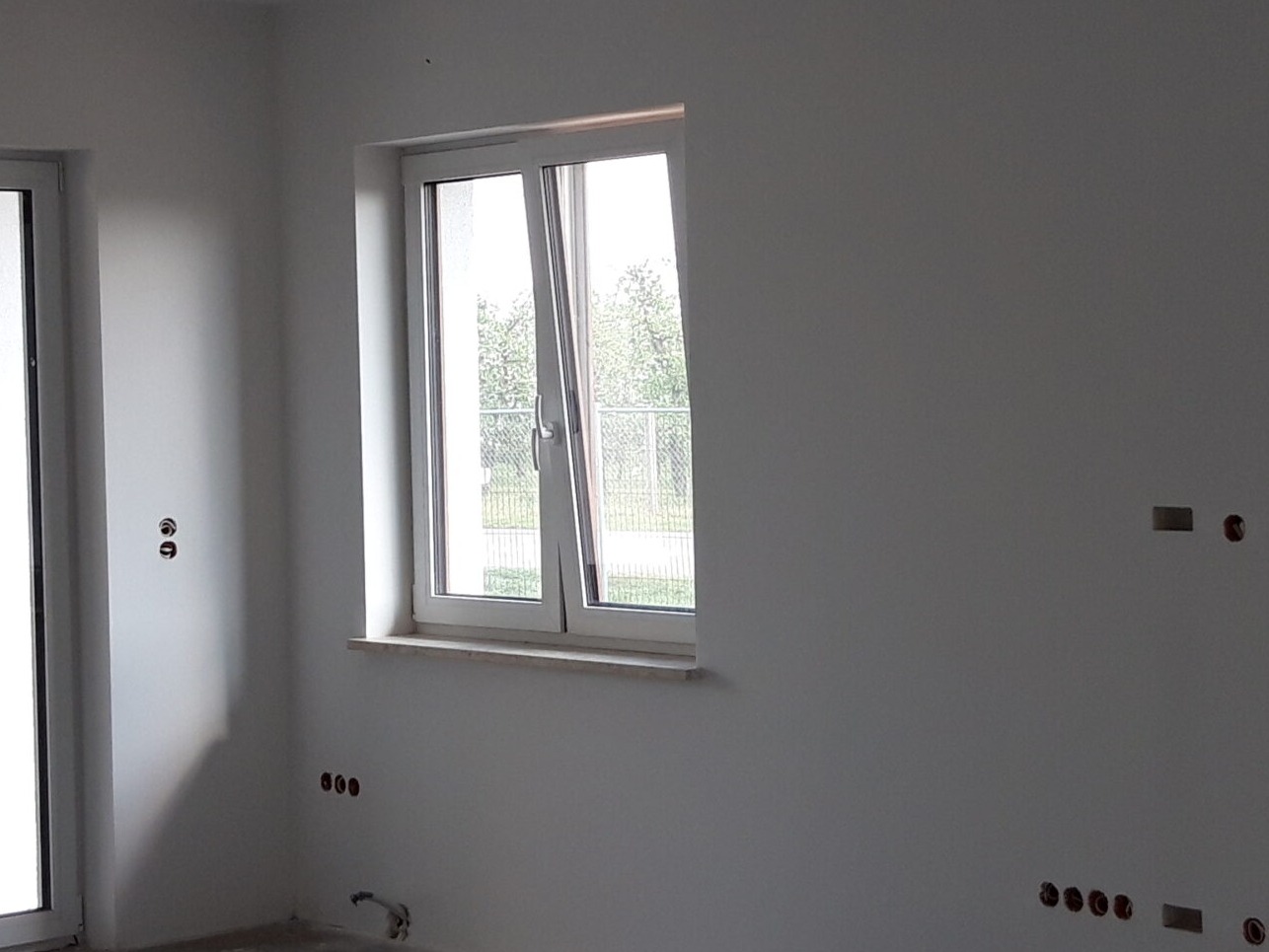 Wnętrze pokoju w stanie deweloperskim z oknem uchylonym, widoczne przygotowane punkty elektryczne w ścianach.