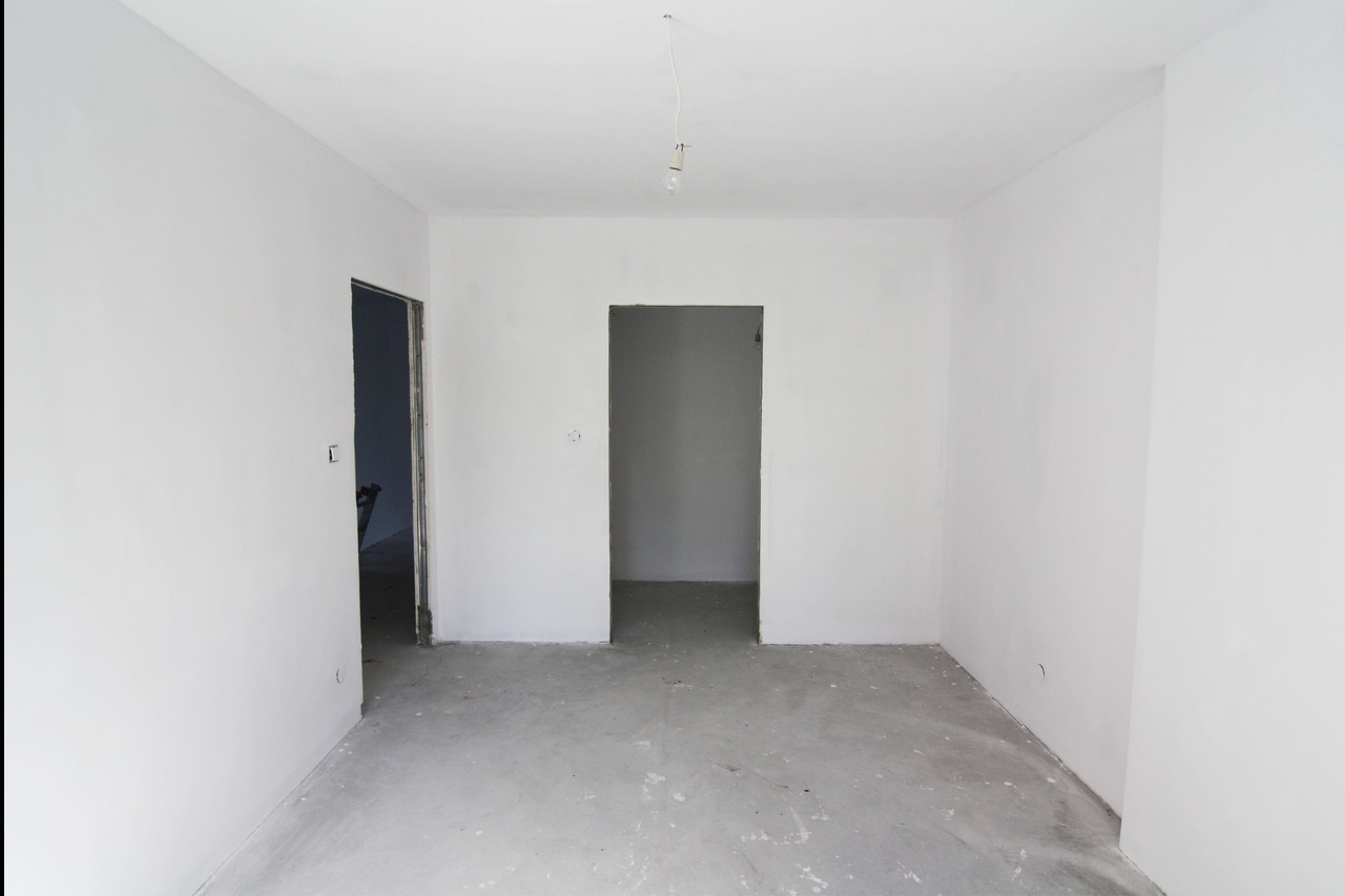 Pusty pokój po remoncie, świeżo wygładzone i pomalowane ściany na biało, surowa podłoga, widoczne dwa otwory drzwiowe.