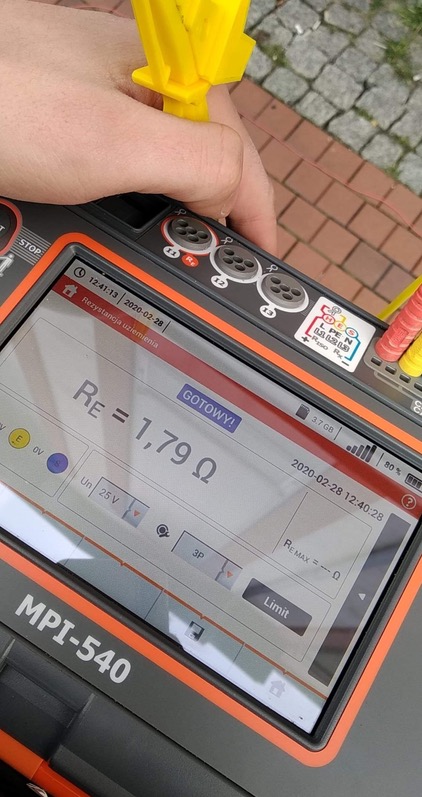 Miernik MPI-540 z wyświetlonym wynikiem pomiaru rezystancji uziemienia 1,79 Ω, trzymany w dłoni na tle brukowanej nawierzchni.