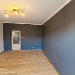 Pomieszczenie po malowaniu: szara ściana kontrastuje z białą, drewniana podłoga, drzwi z szybą i lampa sufitowa. Widok na odnowione wnętrze.