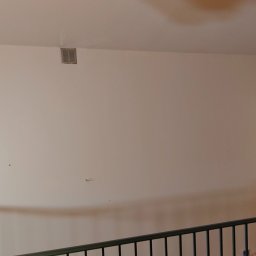 Tomasz Pawlik - Jasna ściana po remoncie z widoczną kratką wentylacyjną pod sufitem i fragmentem balustrady na pierwszym planie. Ciemne drzwi po prawej stronie.