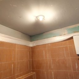 Tomasz Pawlik - Łazienka w trakcie malowania sufitu na szaro, zabezpieczone folią płytki. Widoczne niedomalowane fragmenty i oświetlenie sufitowe. Prace wykończeniowe w toku.