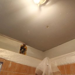 Tomasz Pawlik - Szpachlowana sufitowa powierzchnia z widocznym otworem instalacyjnym, zabezpieczona folią ściana z brązowymi płytkami i oświetlenie sufitowe.