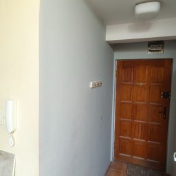 Tomasz Pawlik - Korytarz po odświeżeniu: ściany pomalowane na szaro, widoczny fragment jasnej ściany, brązowe płytki na podłodze i drewniane drzwi wejściowe.