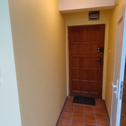 Tomasz Pawlik - Korytarz po odświeżeniu: ściany w ciepłym odcieniu beżu, brązowe płytki na podłodze, drewniane drzwi wejściowe. Widoczne gładkie powierzchnie ścian i sufitu.