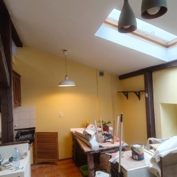 Tomasz Pawlik - Żółte wnętrze w trakcie prac wykończeniowych: widoczne drewniane belki, okno dachowe, narzędzia i materiały malarskie. Przygotowanie do malowania ścian.