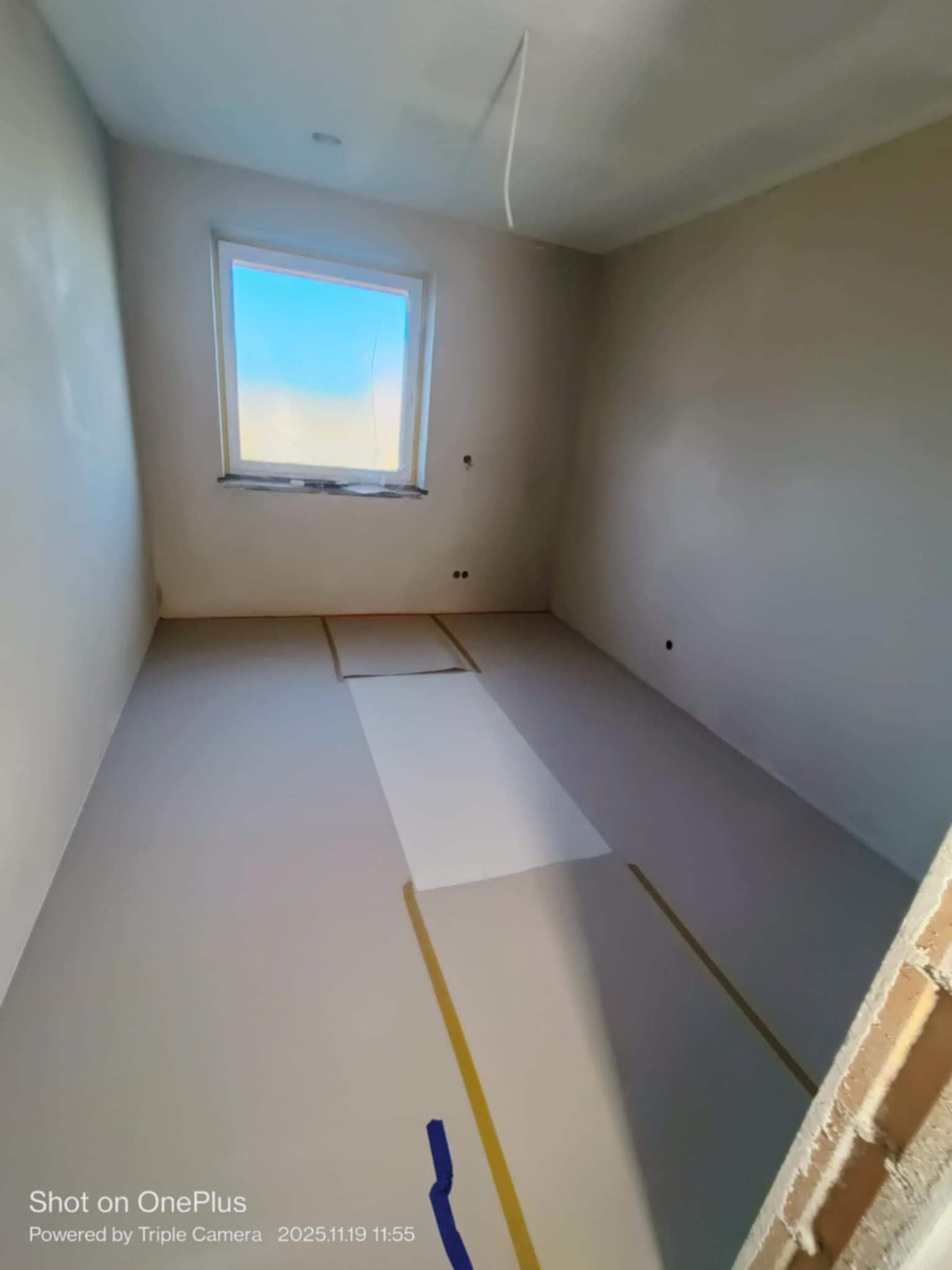 Pokój po szpachlowaniu, przygotowany do malowania. Widoczne zabezpieczenia taśmą malarską okna i podłogi. Ściany gładkie, w jasnym odcieniu szarości. Minimalistyczne wnętrze.