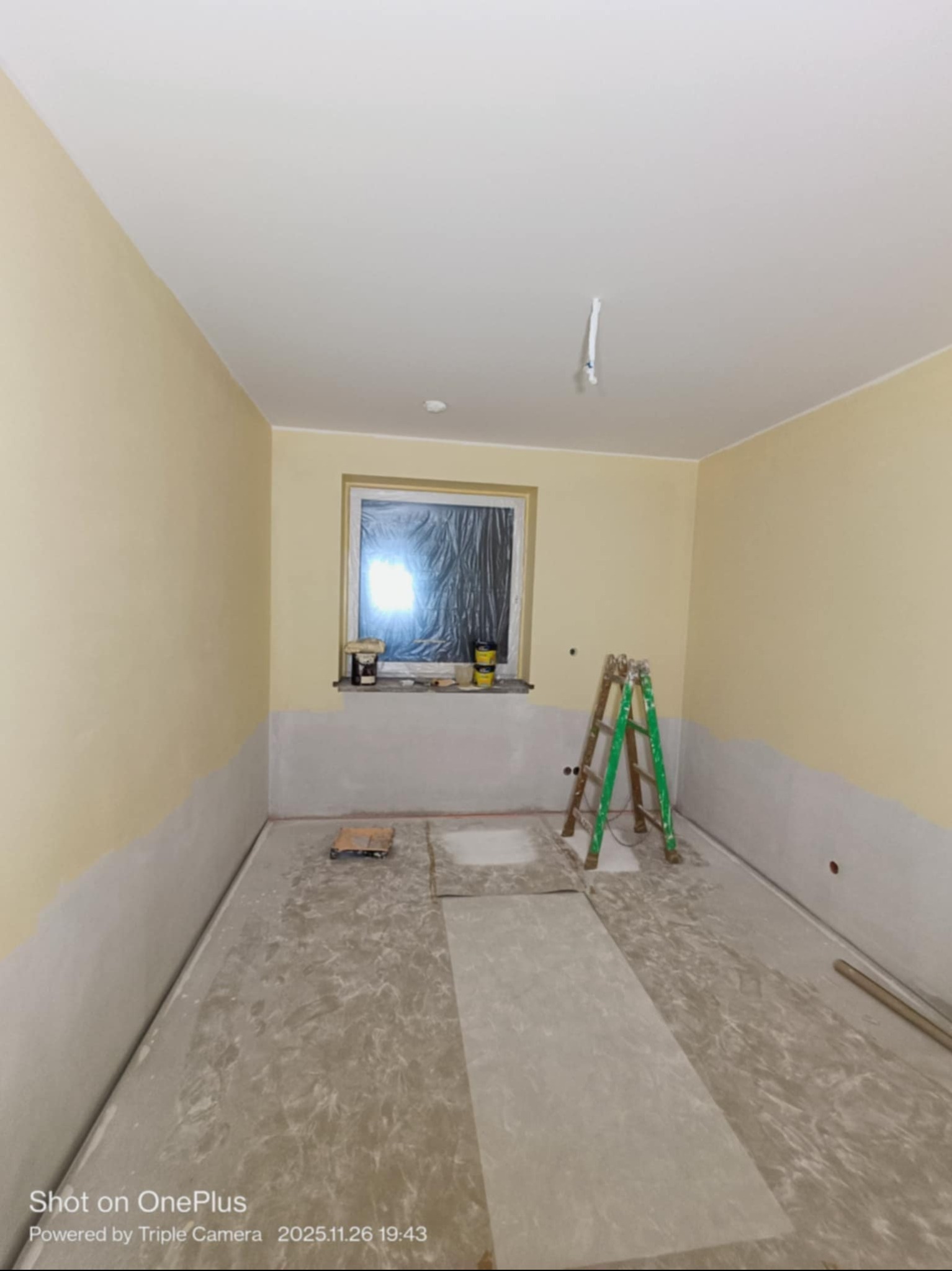 Pokój w trakcie malowania: ściany częściowo pomalowane na żółto, okno zabezpieczone folią, drabina oparta o ścianę. Podłoga zabezpieczona folią malarską.