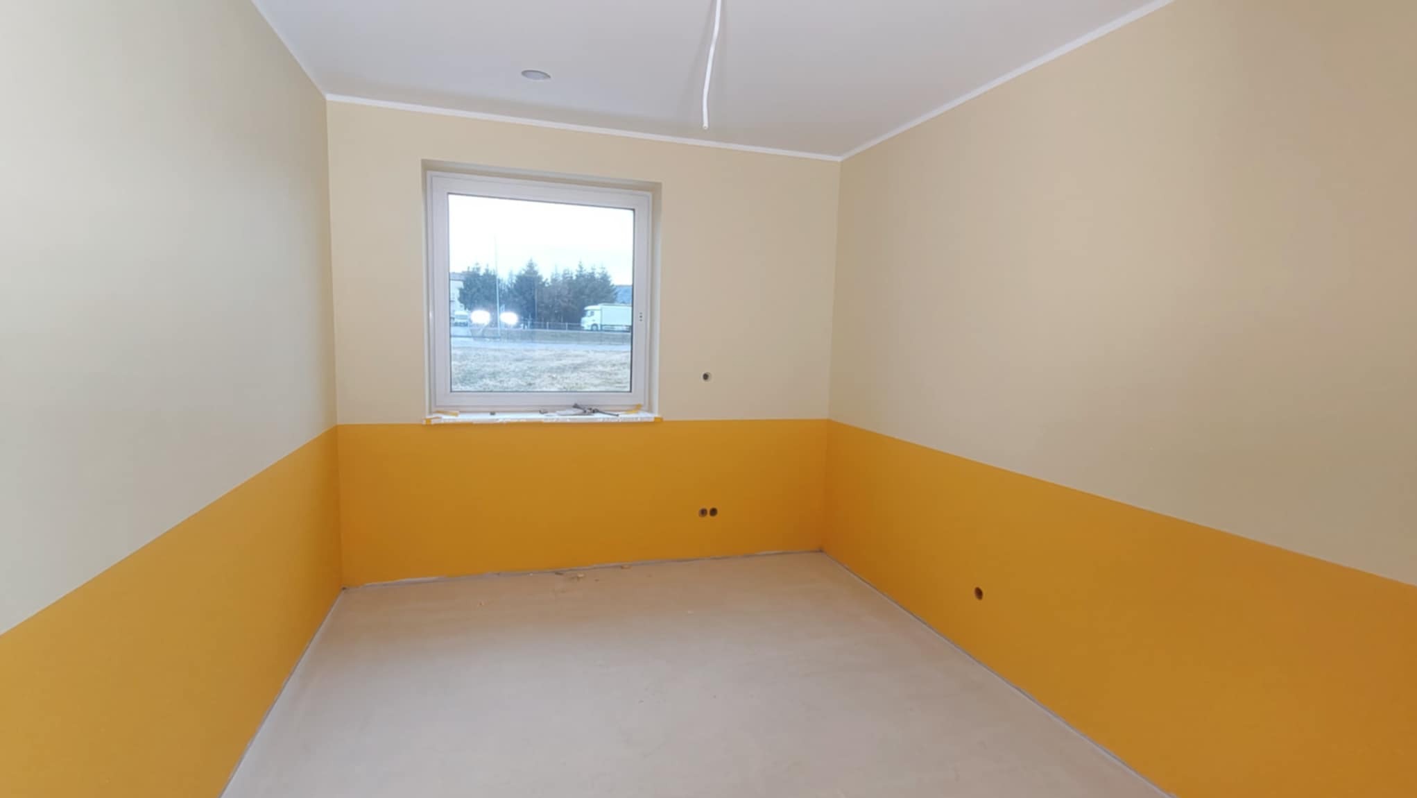 Pokój po częściowym malowaniu ścian na żółto, z widocznymi otworami na gniazdka. Ściany górą w odcieniu beżu, okno z widokiem na zewnątrz. Nowa wylewka na podłodze.