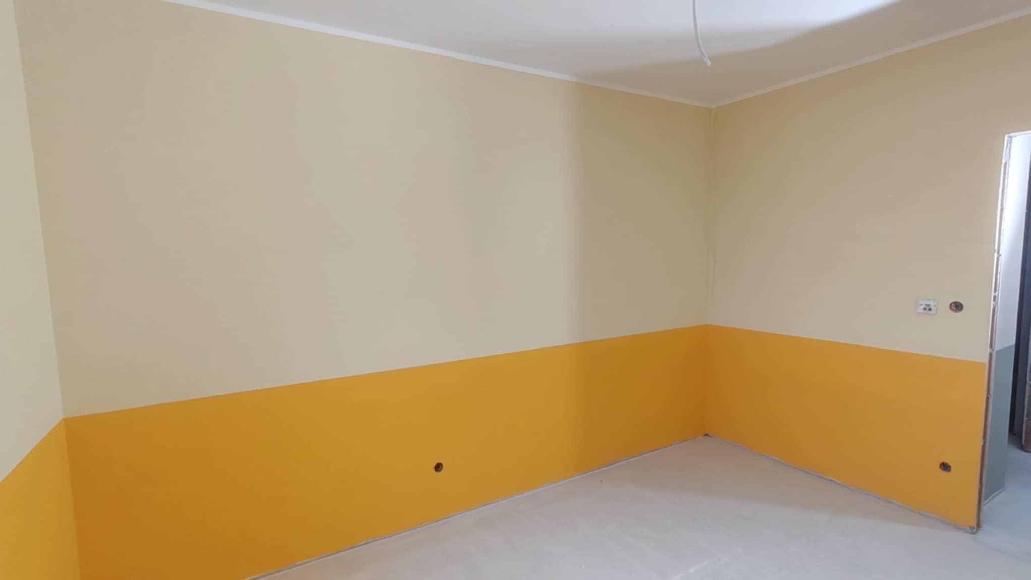 Pokój po malowaniu: ściany podzielone na dwie strefy kolorystyczne - kremowa góra i intensywnie żółty dół. Widoczne gniazdka elektryczne i listwa przypodłogowa.