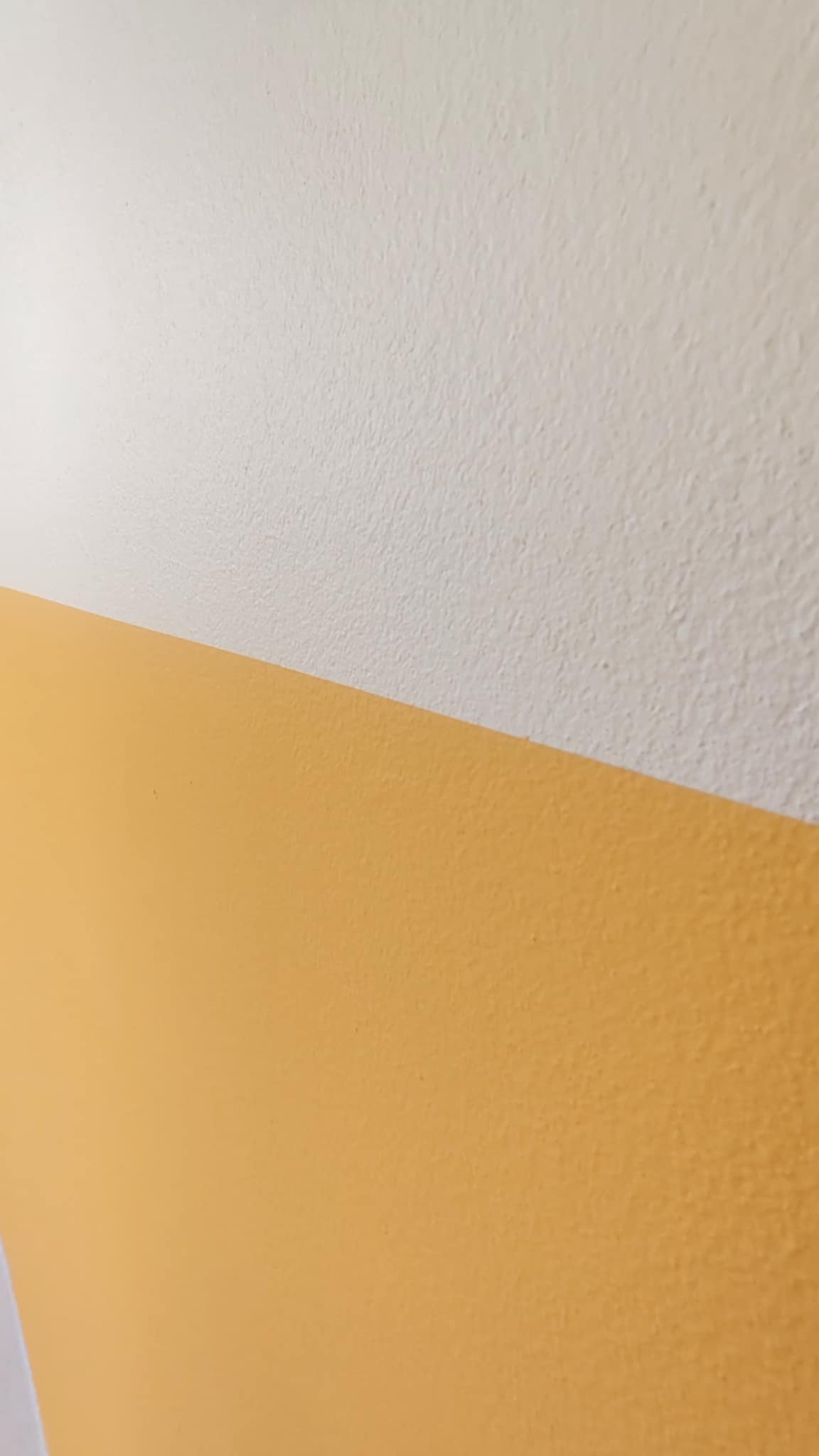 Ściana pomalowana na dwa kolory: kremowy u góry i musztardowy na dole, oddzielone ukośną linią. Widoczna faktura ściany. Ujęcie z dołu, blisko ściany.