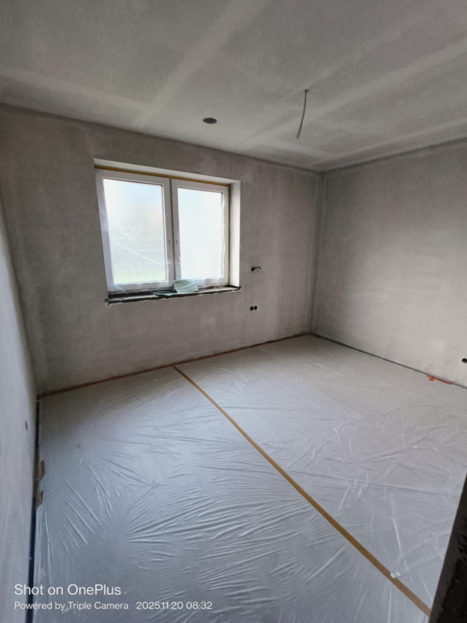 Pokój po szpachlowaniu, przygotowany do malowania. Podłoga zabezpieczona folią, ściany gładkie, okno z parapetem. Widoczne gniazdka elektryczne.