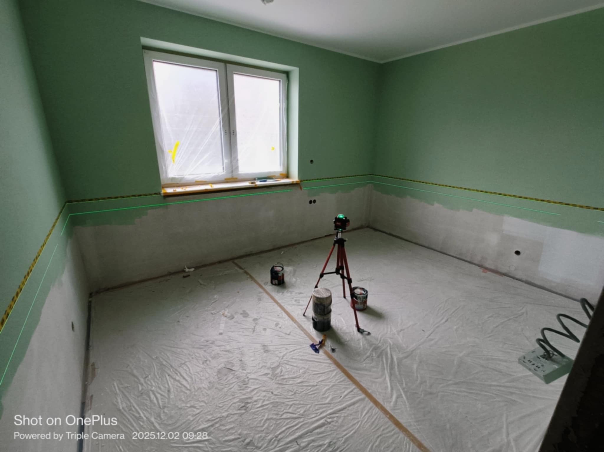 Pokój w trakcie malowania: zielone ściany, dolna część przygotowana, poziomica laserowa, folia ochronna na podłodze, puszki z farbą. Widok na okno.