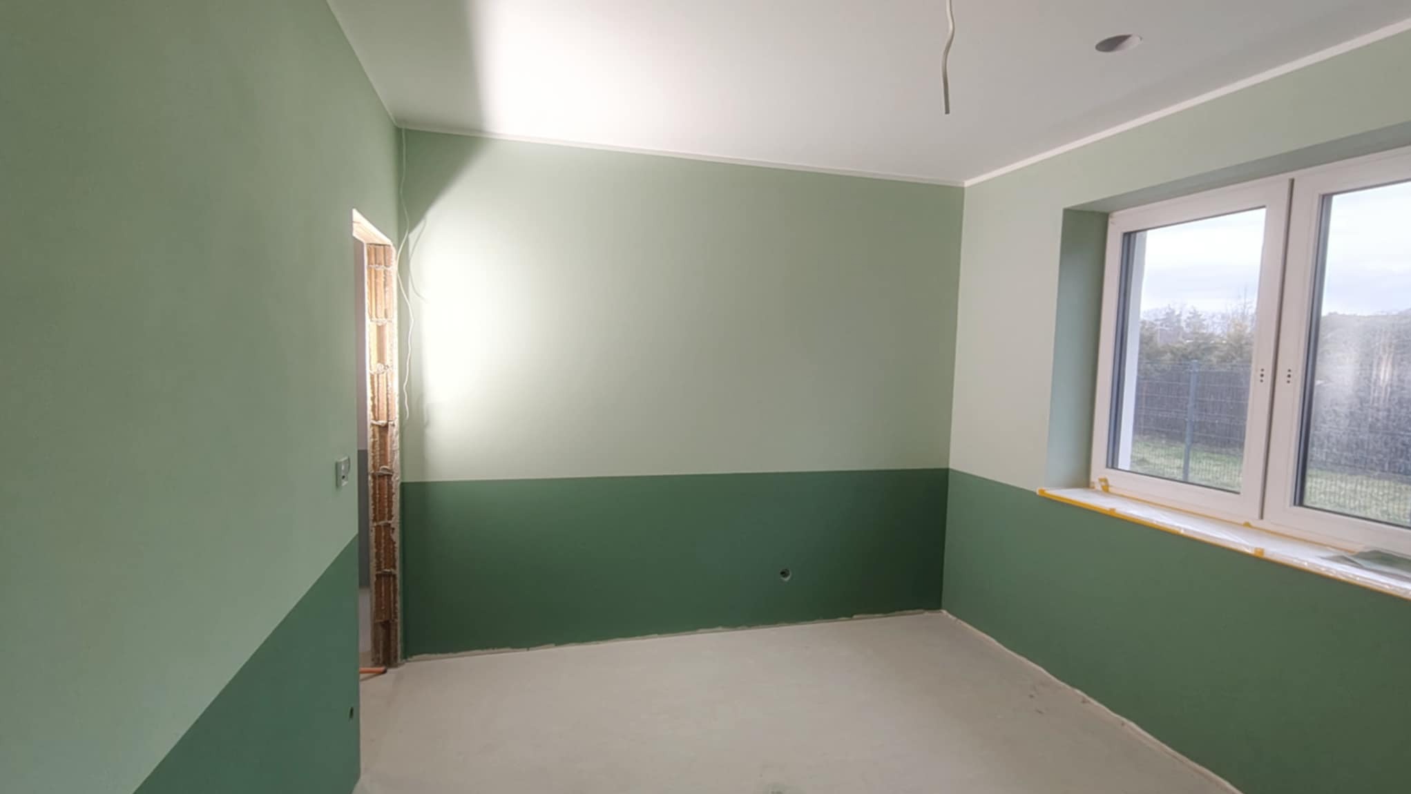 Świeżo pomalowane wnętrze pokoju w odcieniach zieleni, z dwukolorowym podziałem ścian i oknem z widokiem na zewnątrz. Widoczny surowy otwór drzwiowy.