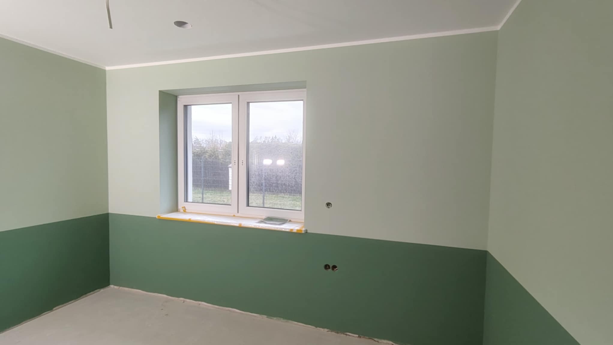 Dwukolorowe ściany w pokoju po malowaniu, dolna część ciemnozielona, górna jasnozielona. Okno z widokiem na zewnątrz. Taśma malarska zabezpiecza parapet.
