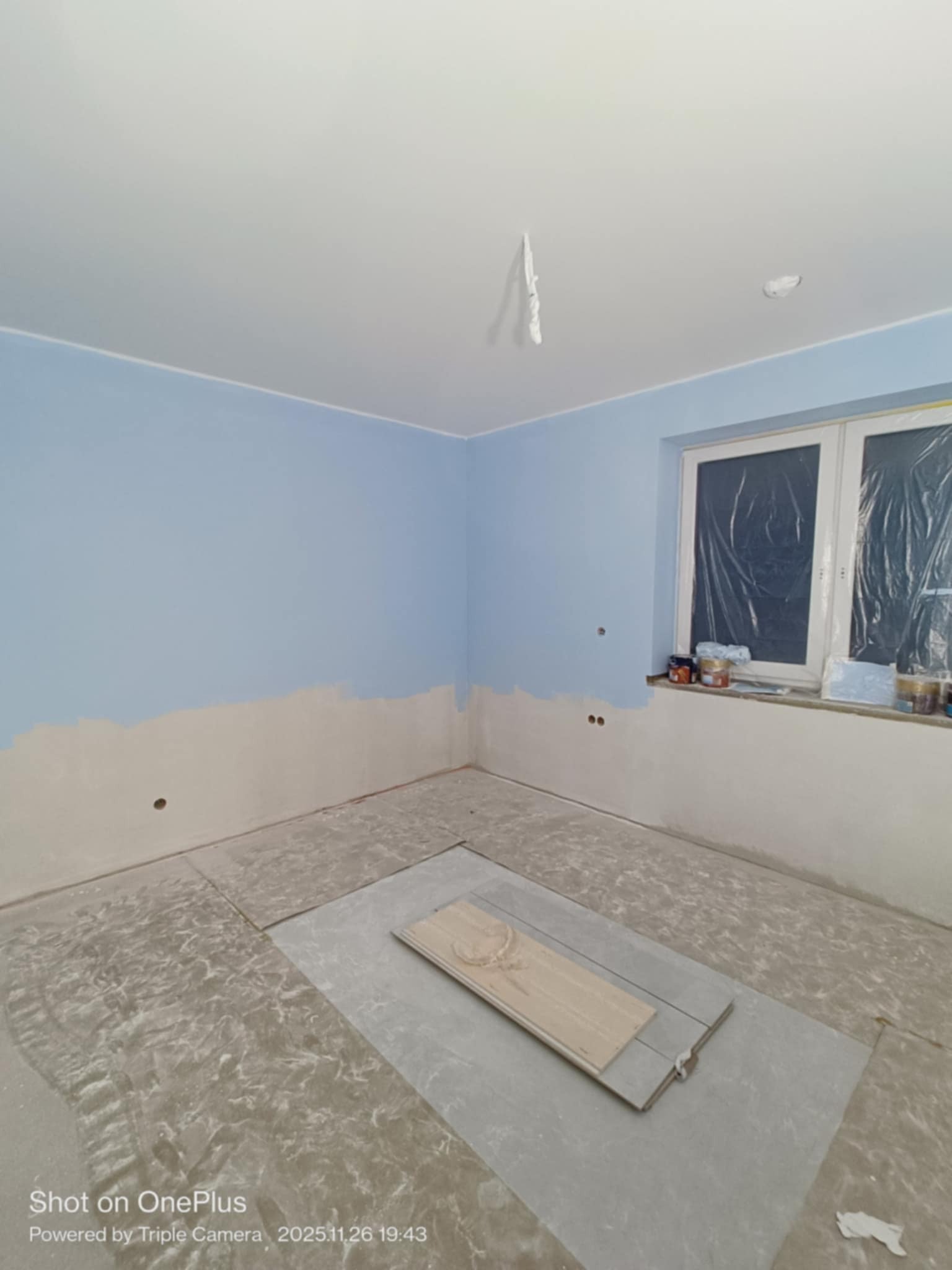 Pokój w trakcie odświeżania w Krakowie: ściany częściowo pomalowane na błękitno, okno zabezpieczone folią, widoczne puszki z farbą. Przygotowanie do dalszych prac.