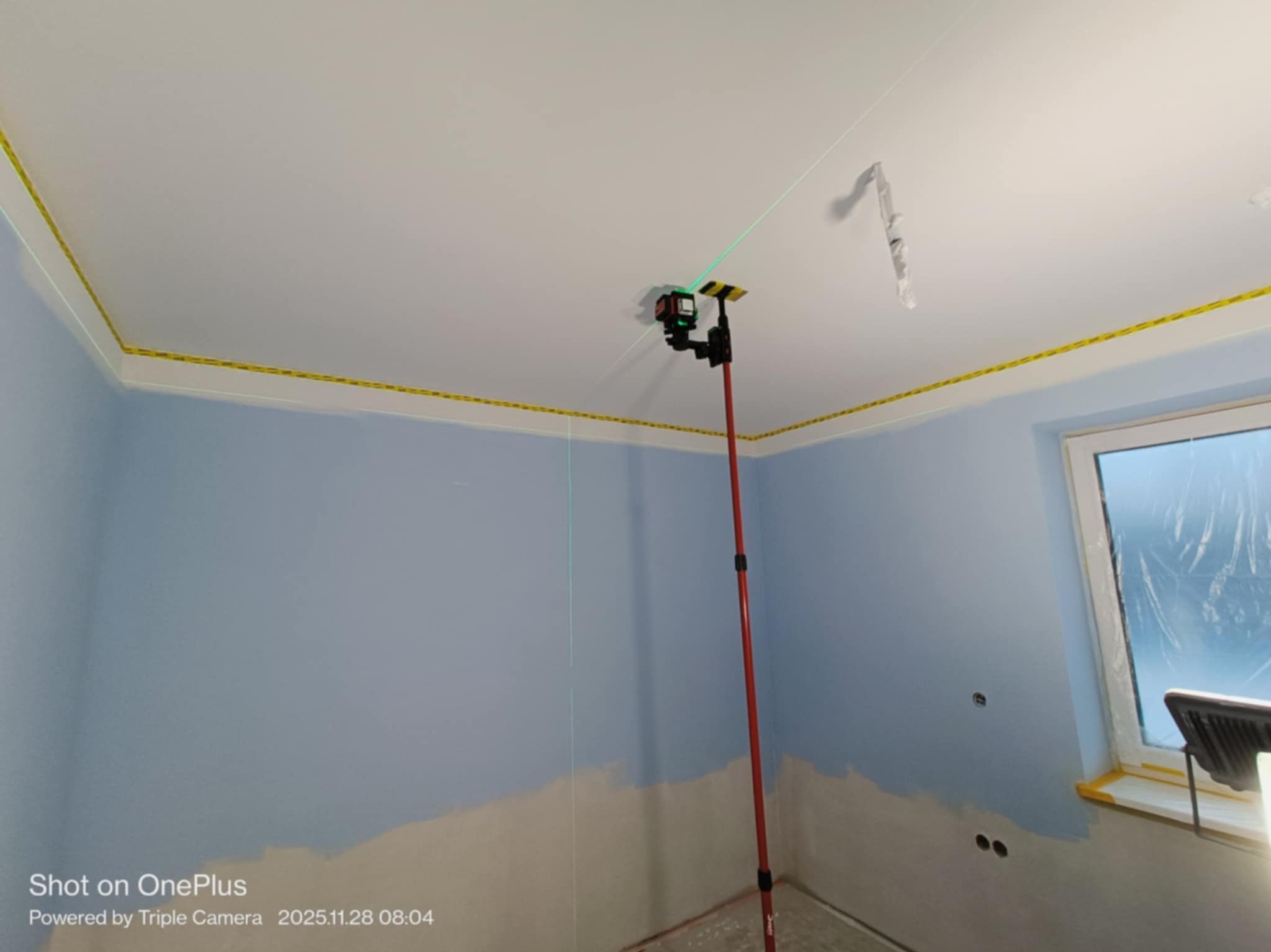 Niebieski pokój w trakcie malowania. Widoczny laser krzyżowy na tyczce, wyznaczający poziom. Ściany częściowo pomalowane, zabezpieczone taśmą malarską.