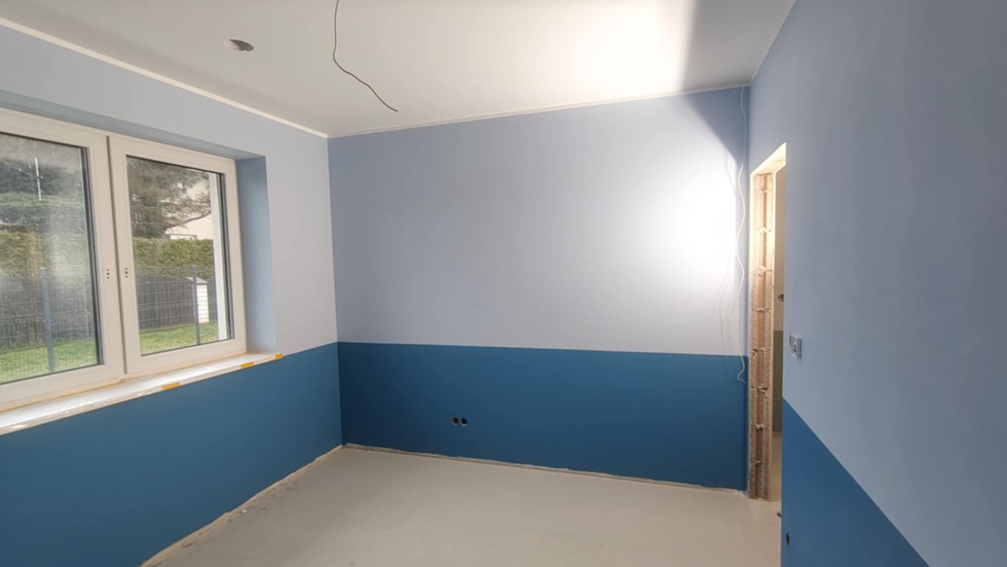 Pomieszczenie po malowaniu ścian na dwa kolory: górna część jasnoniebieska, dolna ciemnoniebieska. Widoczne okno i fragment niezabezpieczonej futryny.