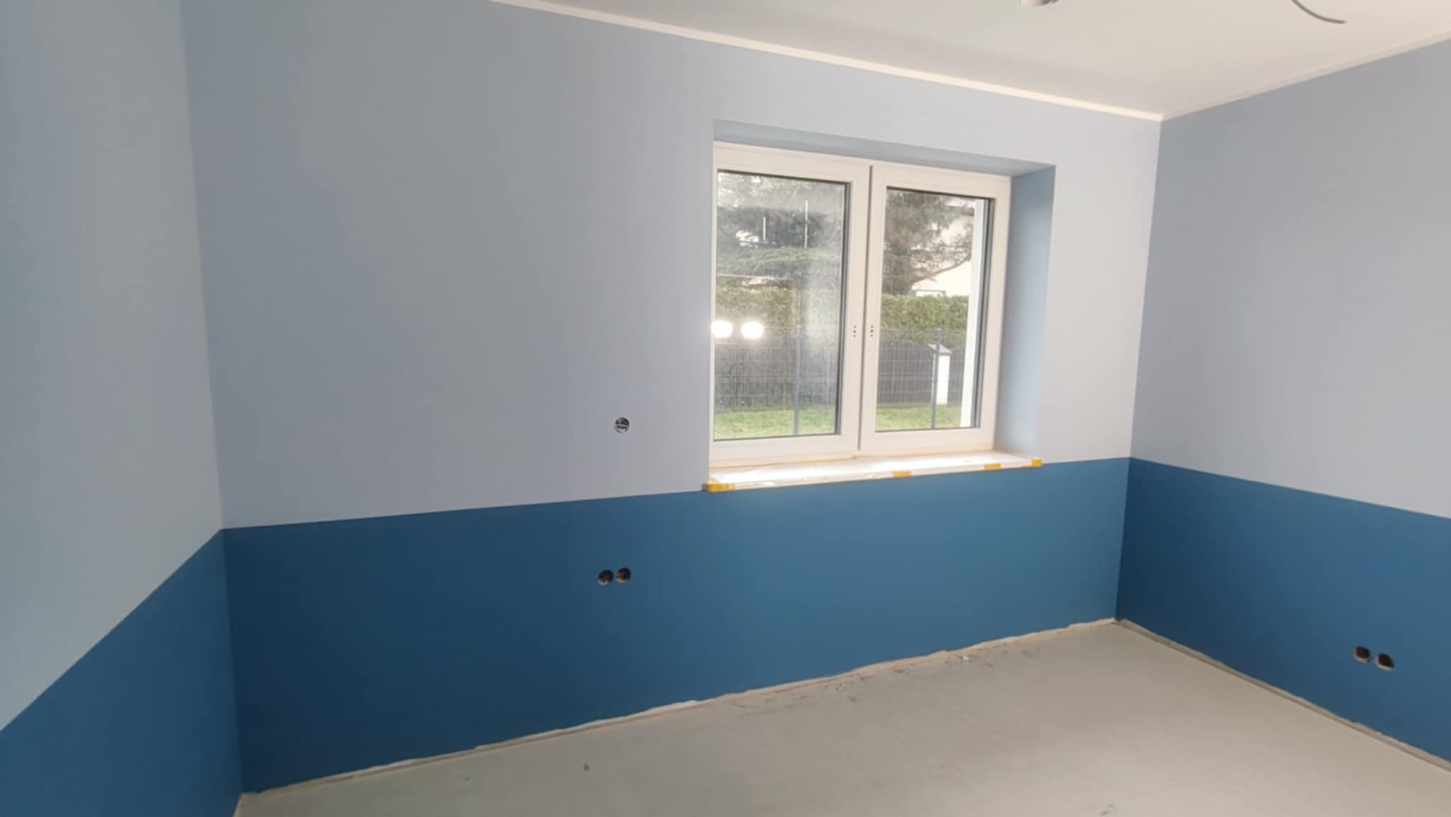 Pokój po malowaniu: ściany dwukolorowe (góra jasnoniebieska, dół ciemnoniebieski), białe okno z żółtą taśmą malarską, szara podłoga, widoczne gniazdka elektryczne.