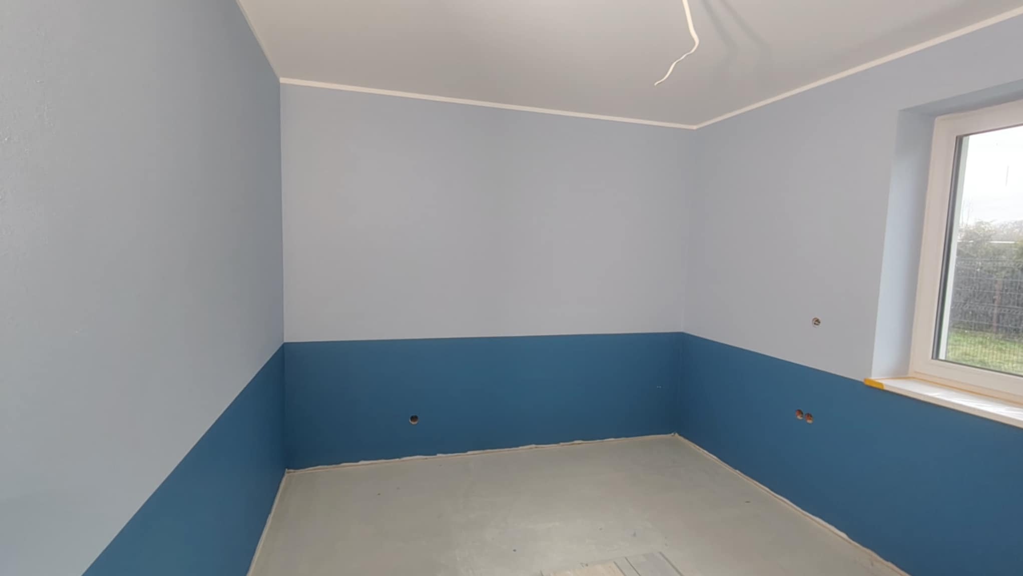 Pomieszczenie po malowaniu: ściany dwukolorowe (góra jasnoniebieska, dół ciemnoniebieski), biały sufit z wystającym kablem, szara podłoga, okno z widokiem na zieleń.