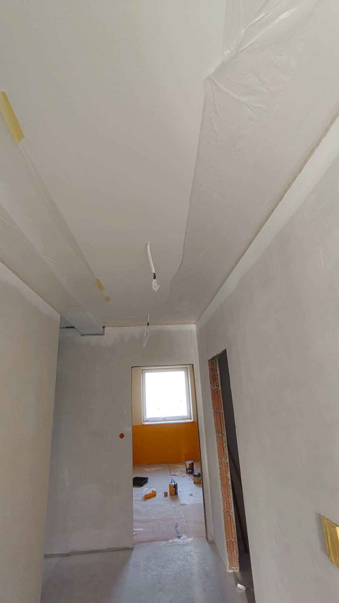 Wnętrze w trakcie prac wykończeniowych: zabezpieczony sufit folią malarską, świeżo wyszpachlowane ściany, widoczne narzędzia i farby w oddali. Przygotowanie do malowania.