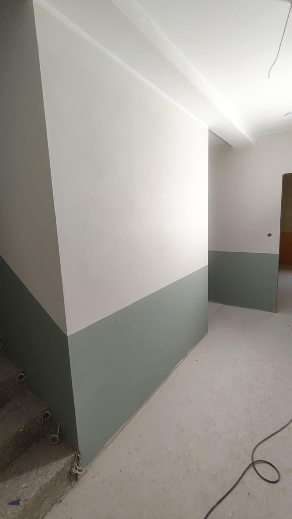Świeżo pomalowane ściany w korytarzu, dolna część w odcieniach szarości, górna biała. Widoczne schody w stanie surowym i wystające kable.