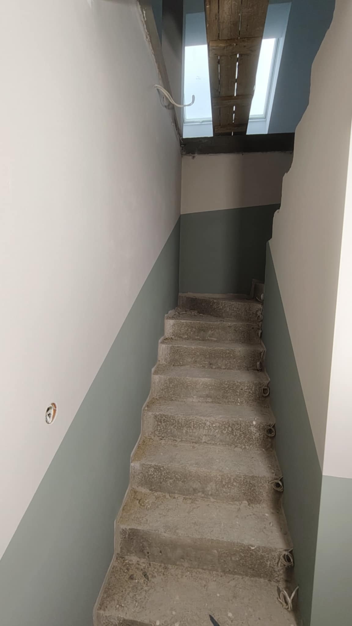 Klatka schodowa w trakcie malowania: ściany pomalowane do połowy na szaro, surowe betonowe schody. Widoczne kable i drewniane rusztowanie przy oknie dachowym.