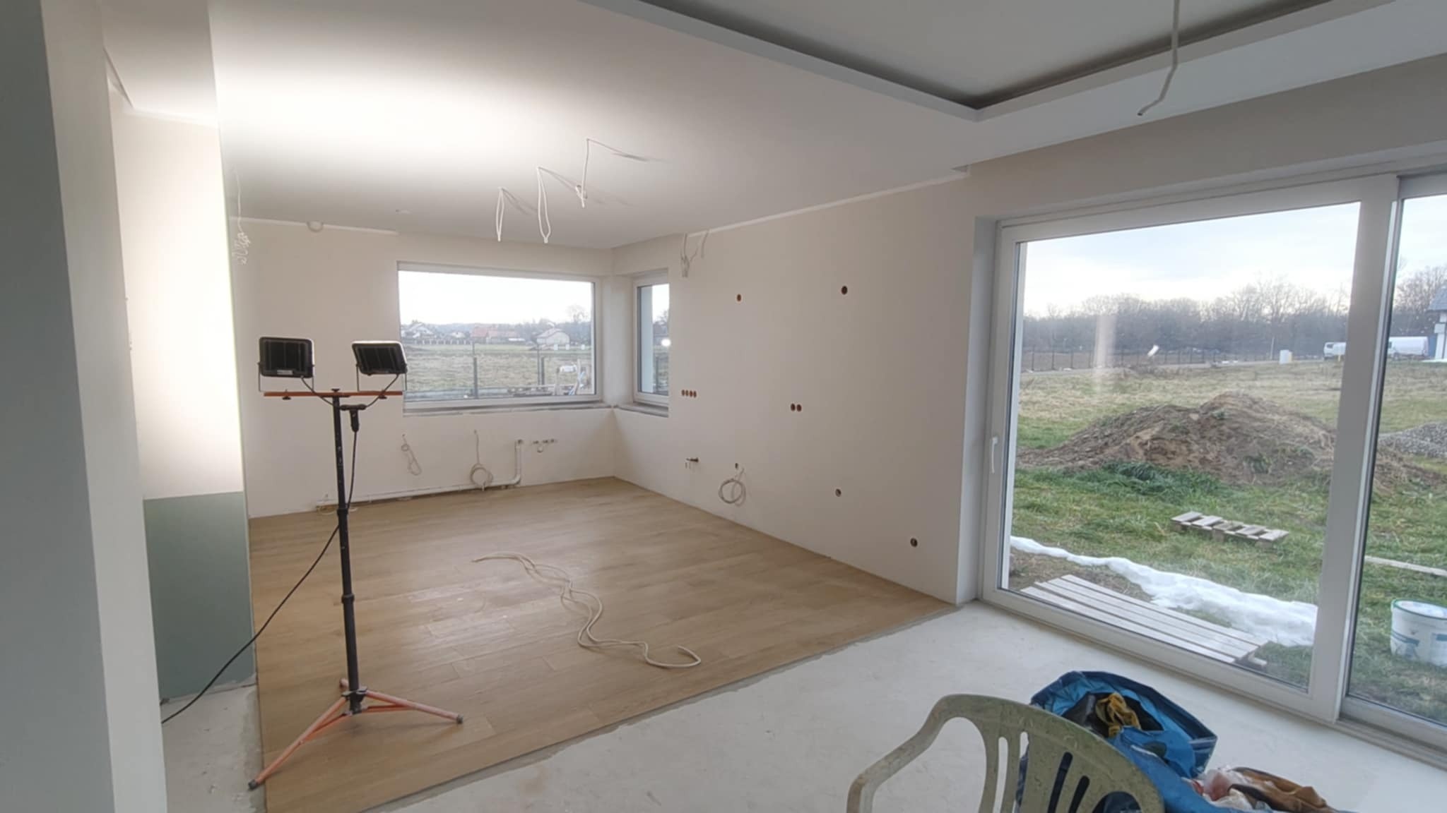 Jasne wnętrze w trakcie prac wykończeniowych: gładkie ściany z widocznymi punktami elektrycznymi, drewnopodobna podłoga, lampa budowlana i widok na zewnątrz.