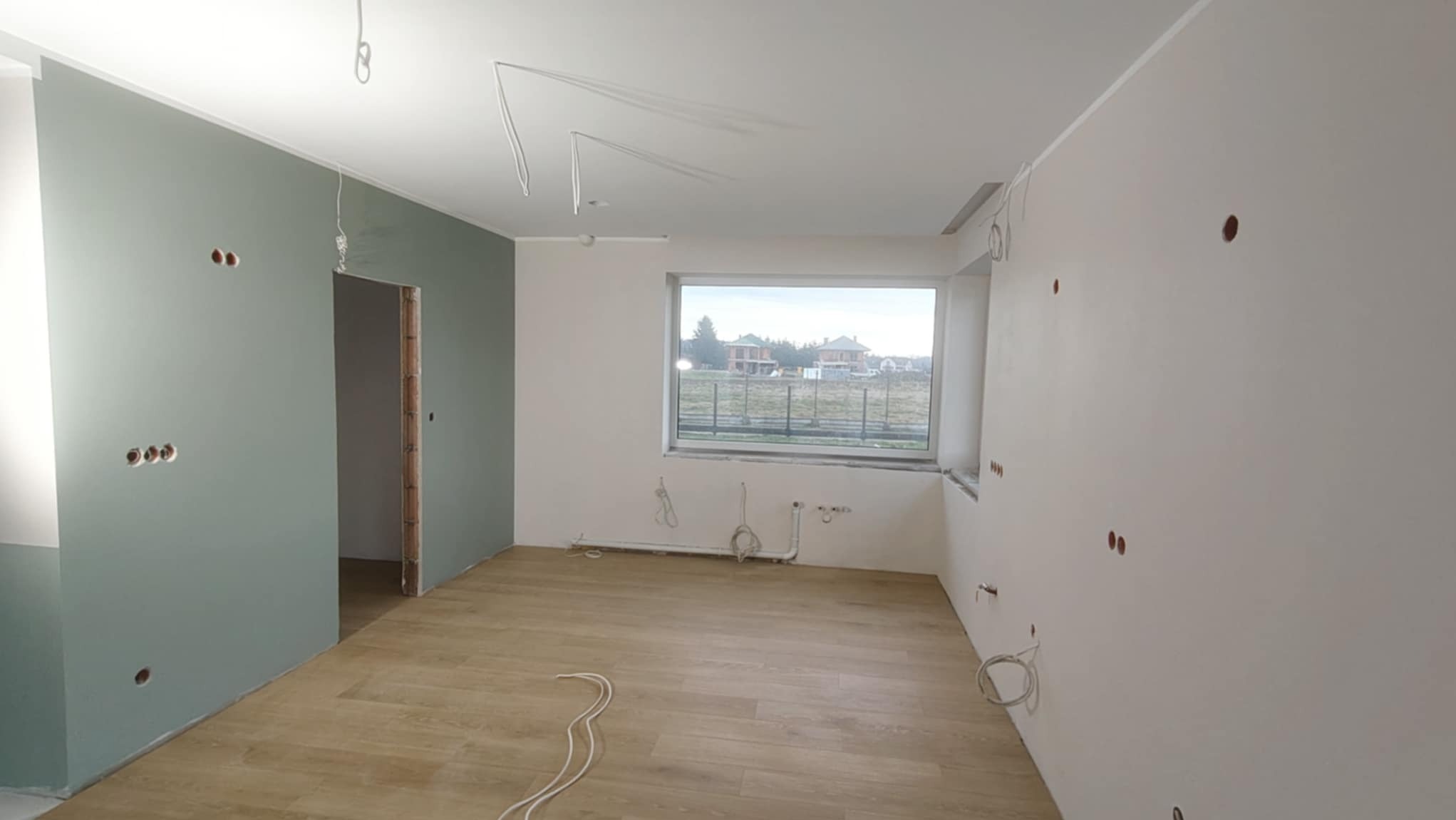Wnętrze po częściowym malowaniu ścian na zielono i biało. Widoczne otwory na gniazdka, okno z widokiem na domy i kable wystające ze ścian i sufitu. Podłoga wyłożona panelami.