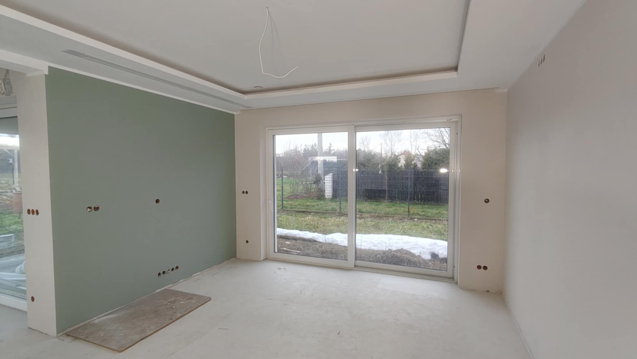 Wnętrze po szpachlowaniu: gładkie ściany w odcieniach bieli i zieleni, widoczne puszki elektryczne, duże okno przesuwne z widokiem na ogród.