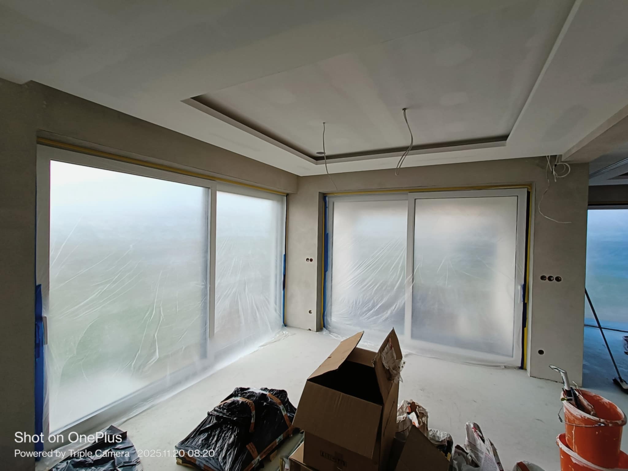 Pokój w trakcie prac wykończeniowych: okna zabezpieczone folią, ściany przygotowane do malowania, widoczne narzędzia i materiały malarskie. Sufit z podwieszaną konstrukcją.