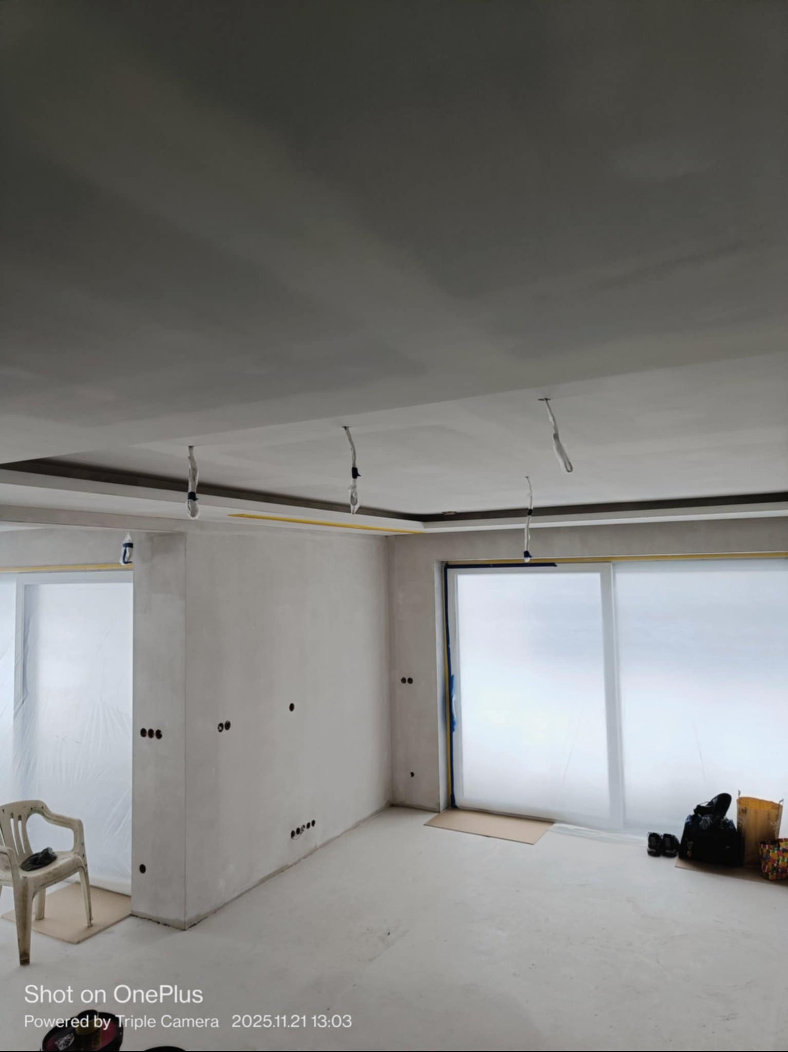 Wnętrze w trakcie szpachlowania: gładkie, białe ściany, zabezpieczone okna folią, widoczne puszki elektryczne. Przy ścianie stoi krzesło.