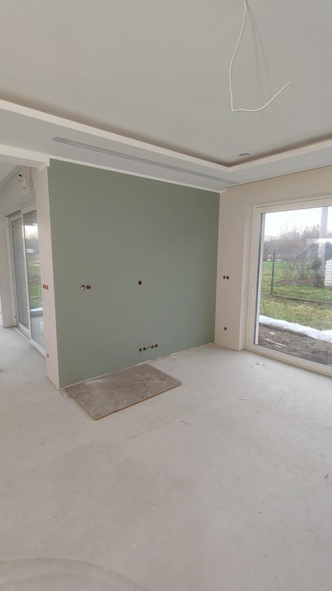 Wnętrze po malowaniu: ściana w odcieniach zieleni z widocznymi otworami na gniazdka, surowa podłoga, okno z widokiem na zewnątrz. Minimalistyczny design, etap wykończeniowy.