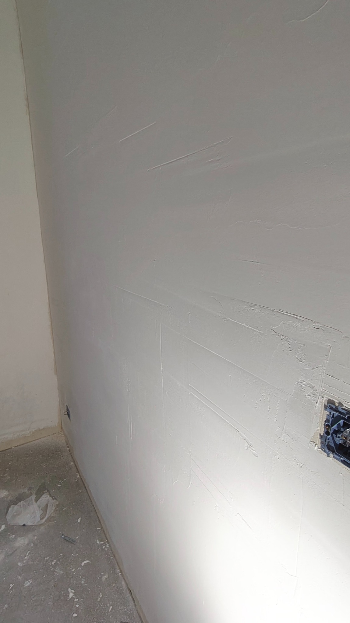 Ściana po nałożeniu gładzi w trakcie prac wykończeniowych w Krakowie. Widoczne ślady po szpachlowaniu i puszka elektryczna. Jasne, naturalne oświetlenie.