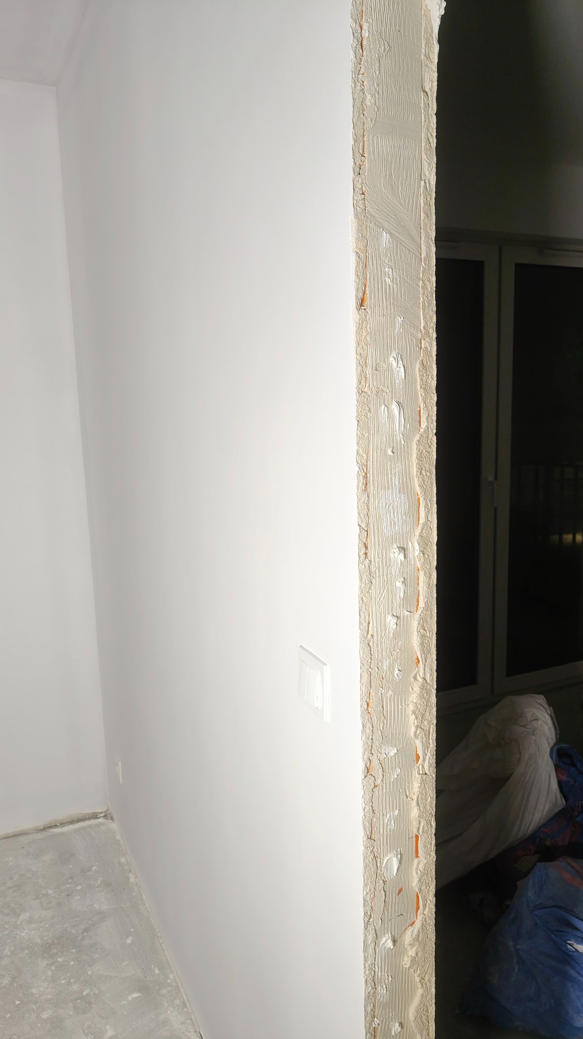 Biała ściana po szpachlowaniu, widoczna krawędź ościeżnicy z nałożoną warstwą szpachli. W tle okno i zakryte folią meble. Minimalistyczne ujęcie wnętrza w trakcie remontu.