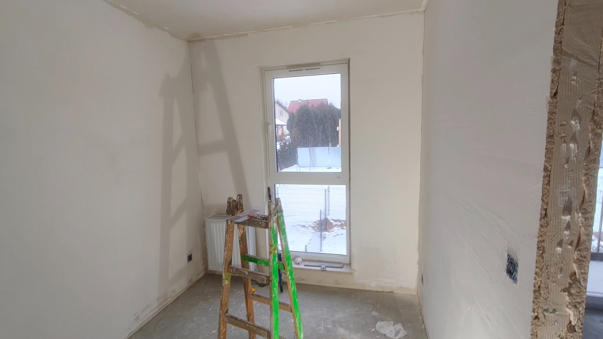 Pokój w trakcie prac wykończeniowych: ściany po szpachlowaniu, drabina malarska, widok na zewnątrz przez okno. Przygotowanie do malowania.