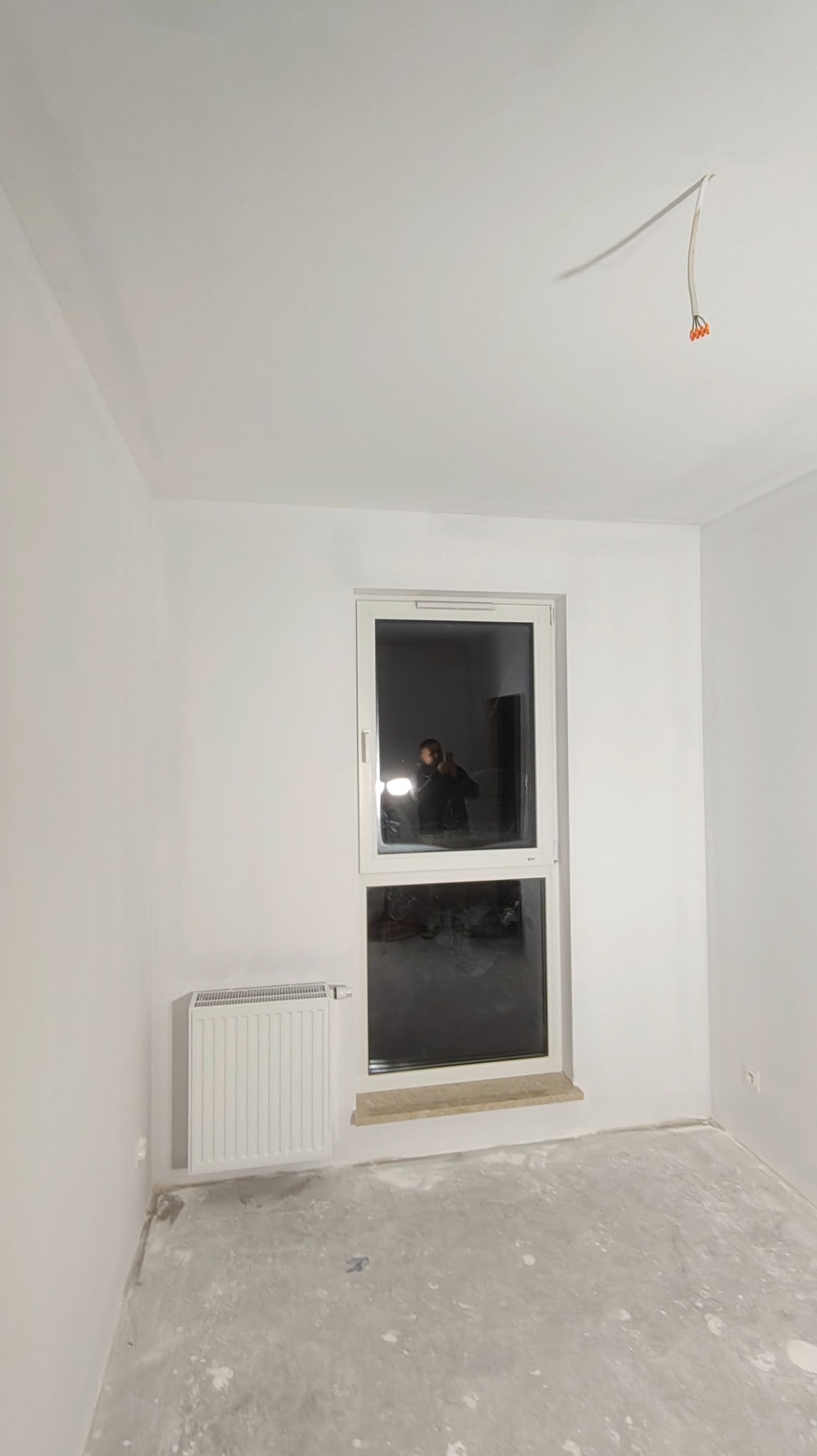Białe, świeżo pomalowane wnętrze pokoju z oknem, grzejnikiem i wystającymi kablami z sufitu. Na podłodze widoczne ślady po pracach wykończeniowych.