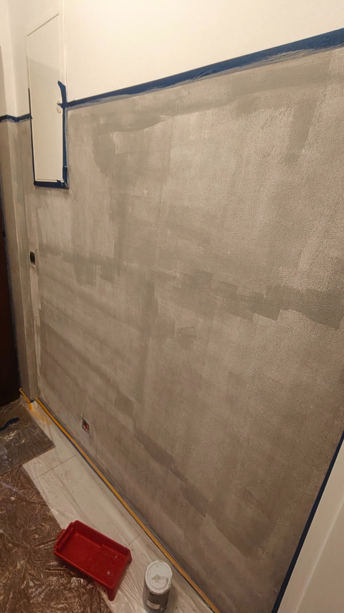 Ściana w trakcie malowania na szaro, zabezpieczona taśmą malarską przy suficie i podłodze. Widoczna kuweta i puszka z farbą. Etap prac wykończeniowych.