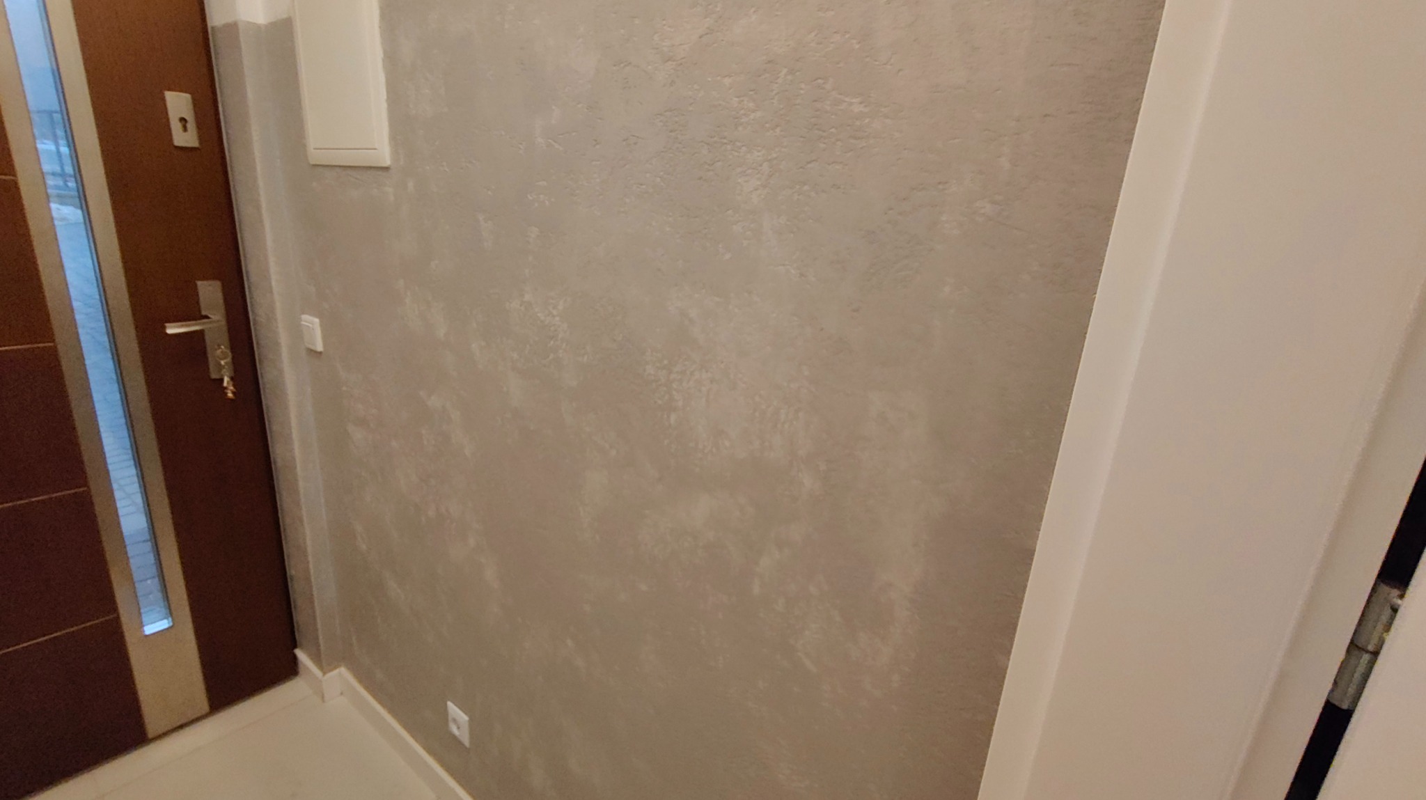 Ściana z efektem betonu dekoracyjnego w korytarzu, widoczna część drzwi wejściowych i biała listwa przypodłogowa. Neutralna kolorystyka, delikatna faktura.